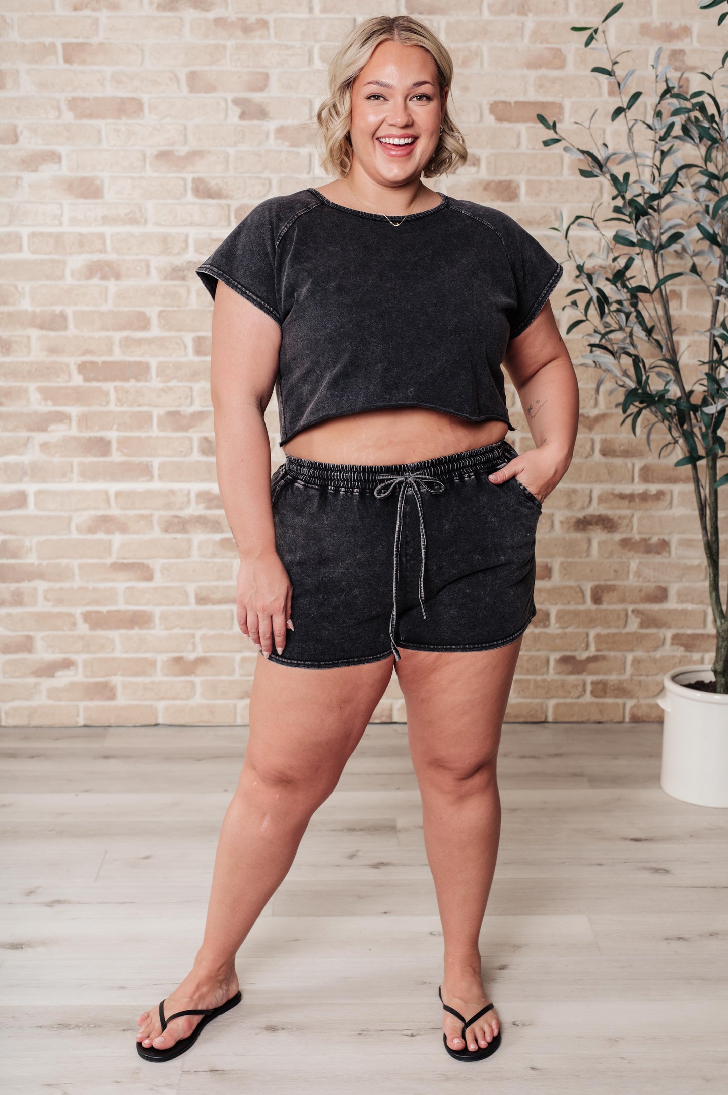 Woman wearing a black t-shirt and shorts standing against a brick wall.