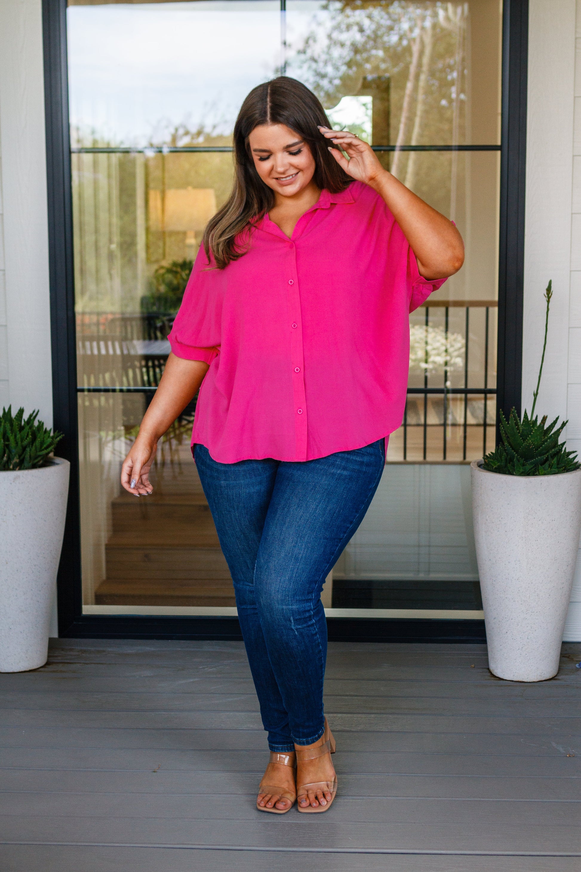 Woman wearing a bright pink blouse and blue jeans standing on a porch.
