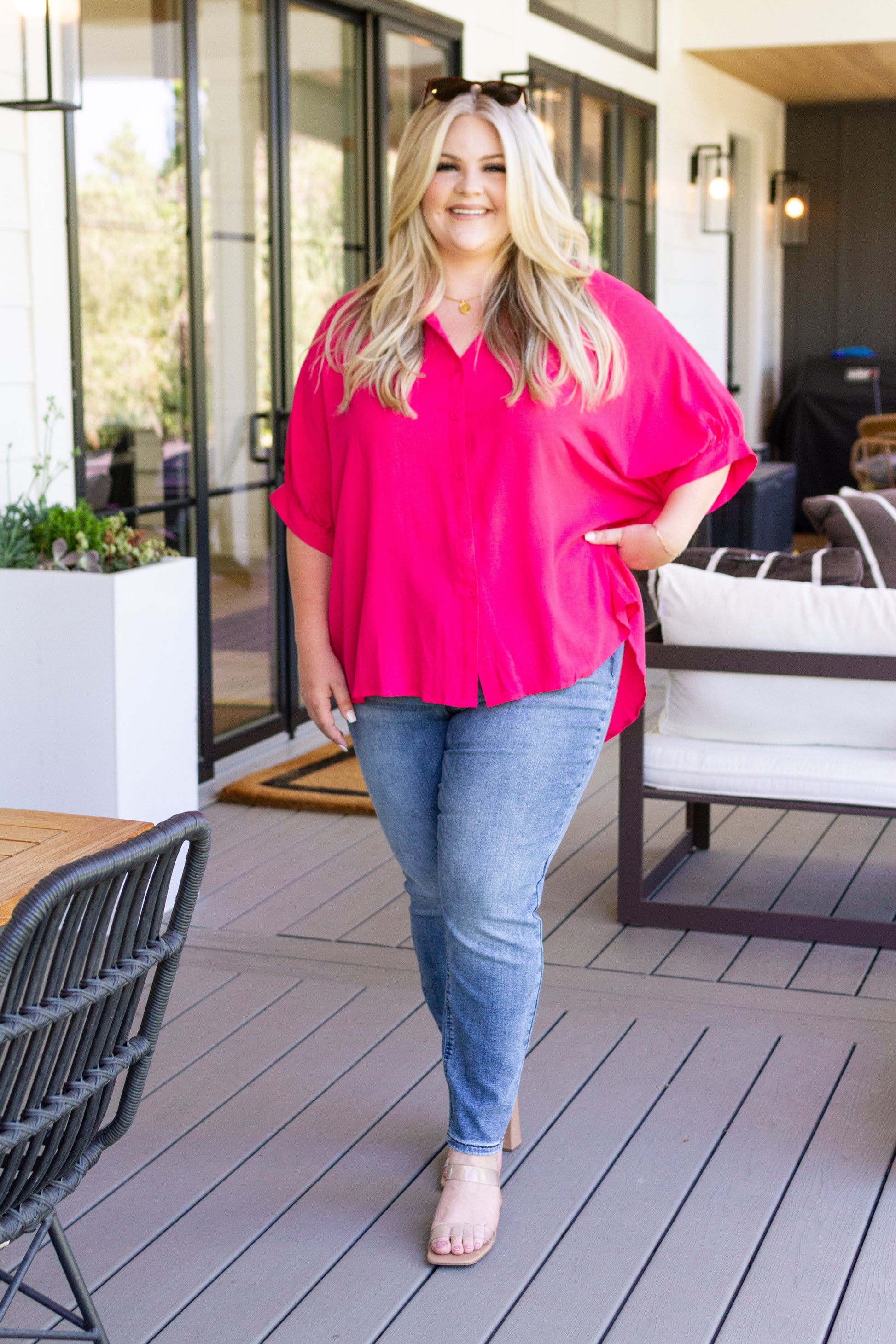 Woman in a bright pink blouse and blue jeans standing on a patio.