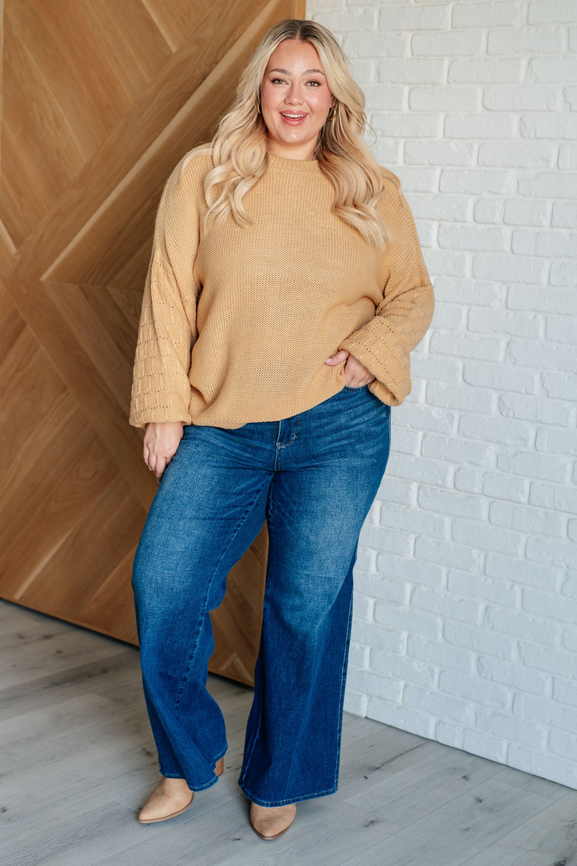 Woman wearing a mustard yellow sweater and blue jeans standing against a white brick wall.