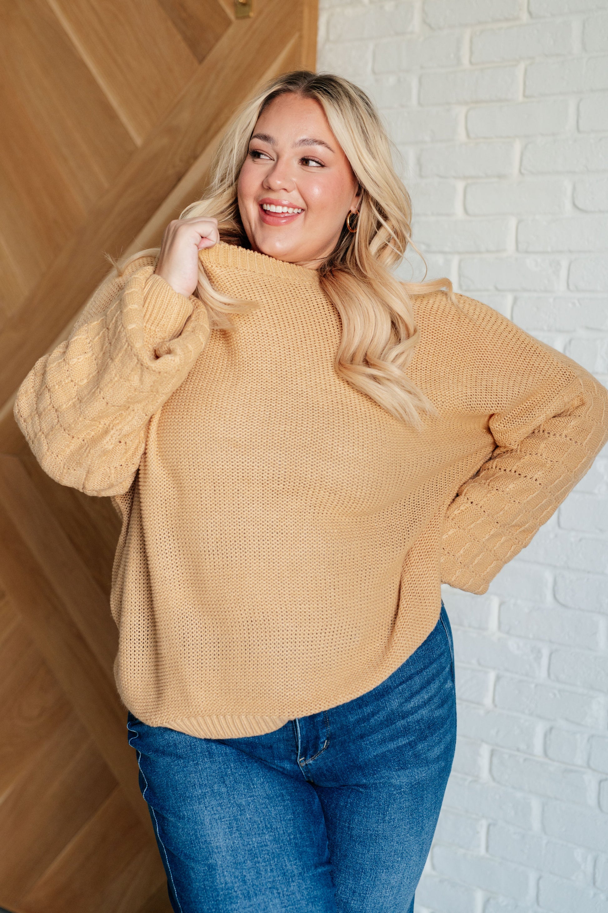 Woman wearing a mustard yellow sweater and blue jeans standing against a white brick wall.