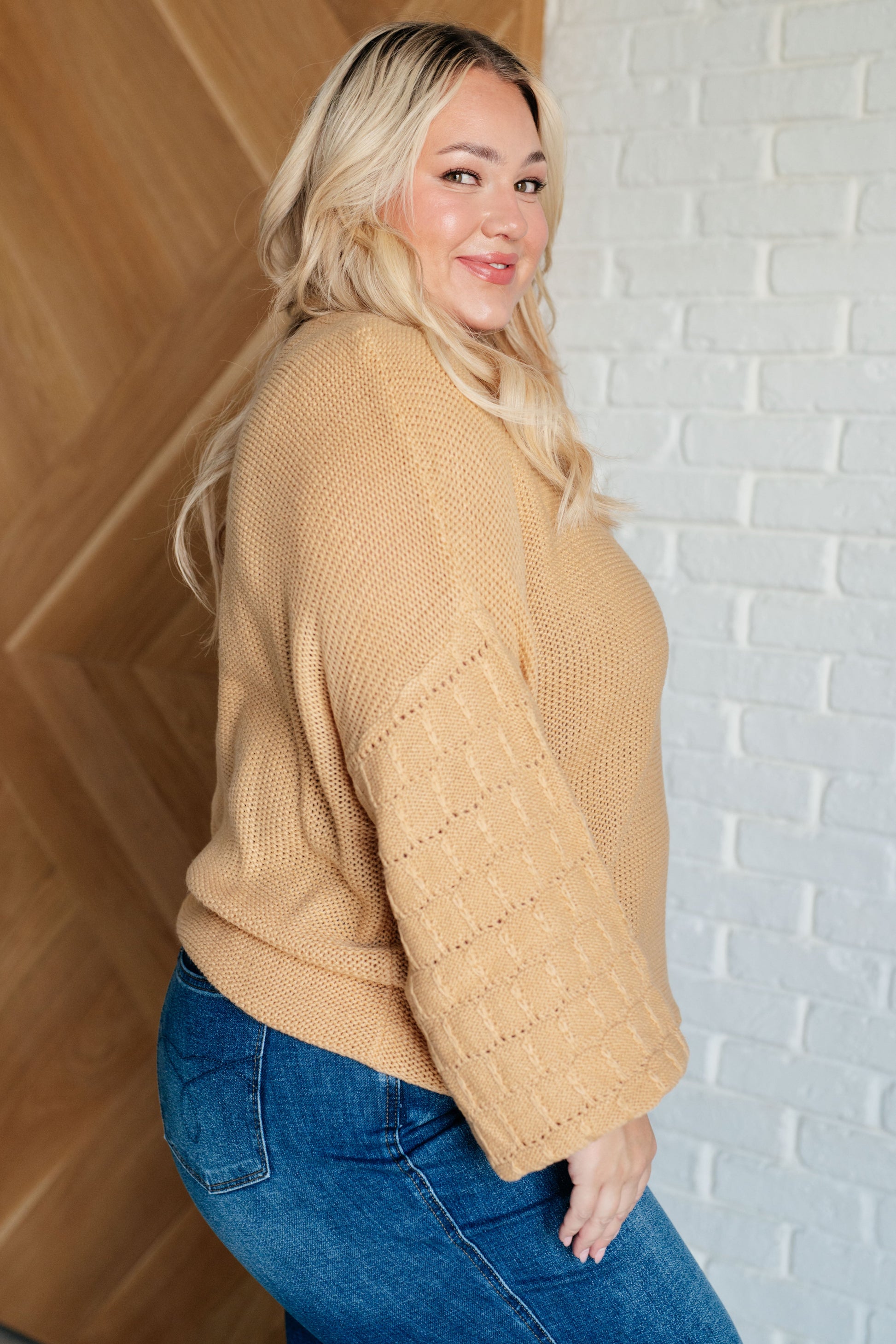 Woman wearing a beige sweater and blue jeans against a white brick wall.