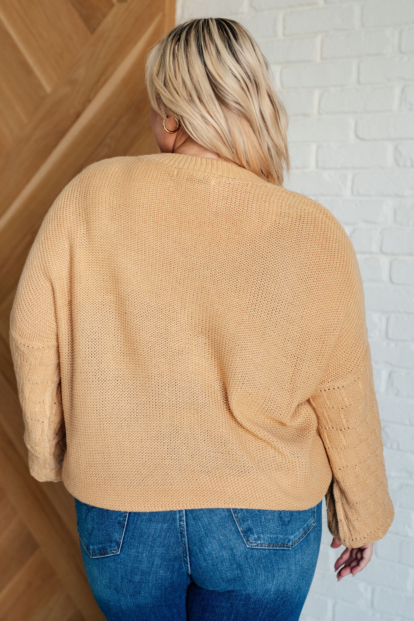 Person wearing a beige sweater and blue jeans against a white brick wall.