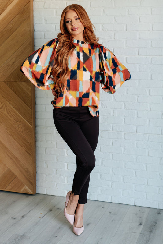 Woman wearing a colorful checkered blouse and black pants against a white brick wall.