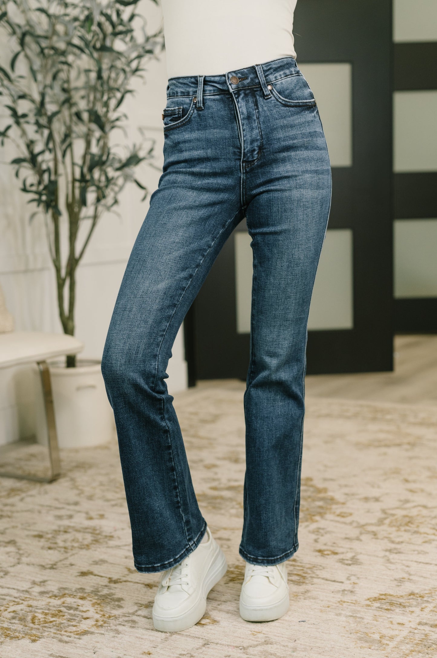A person wearing a pair of medium-wash blue denim jeans with white sneakers, standing on a patterned beige rug in an indoor setting with a potted plant and dark paneled wall in the background.