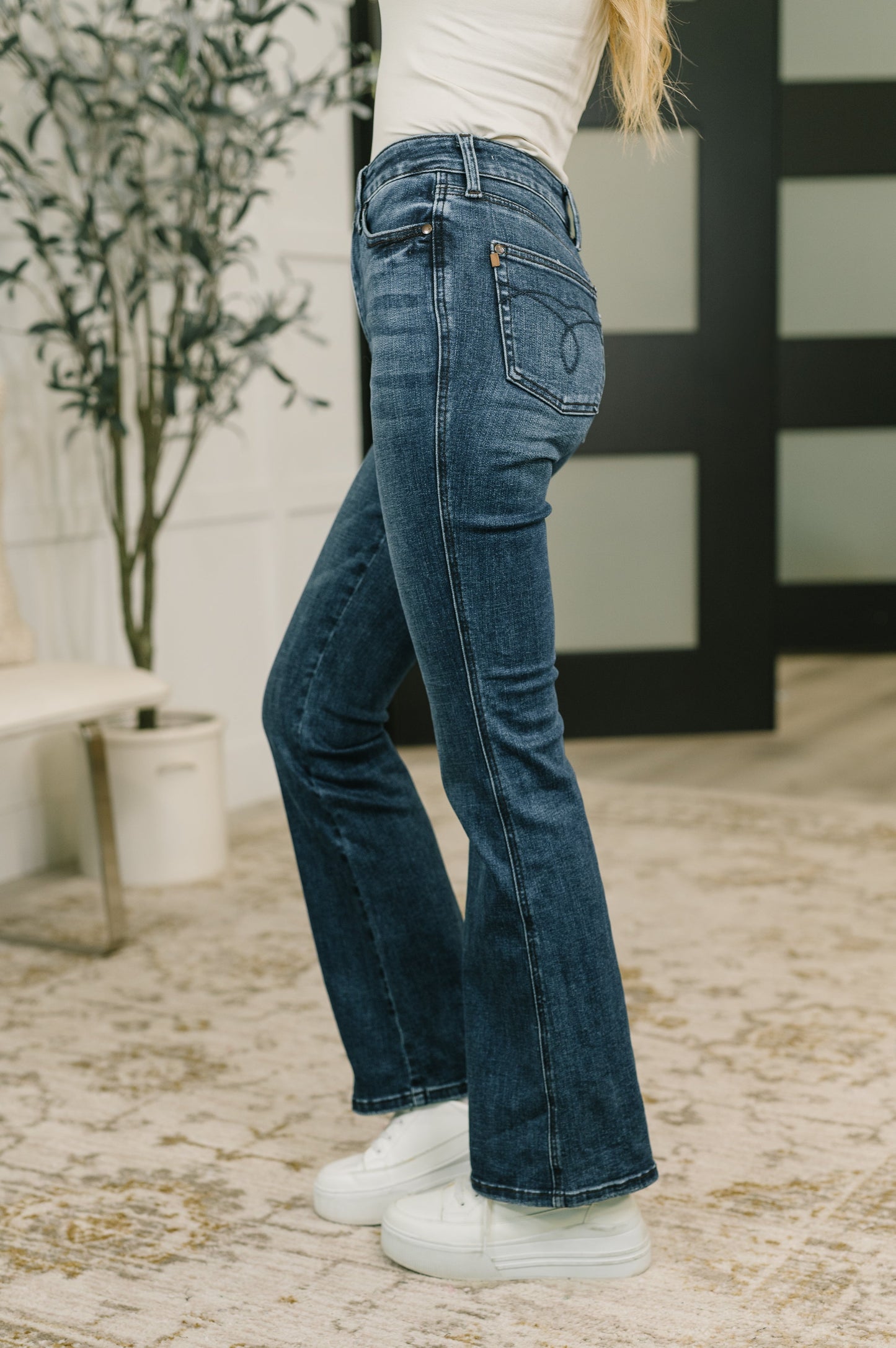 A person wearing dark blue flare jeans, a white top, and white sneakers, standing on a patterned rug in an indoor setting.