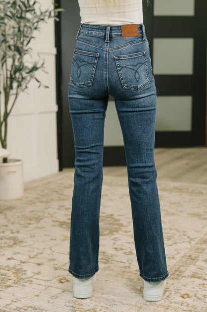 Rear view of a person wearing medium-wash blue flare jeans with white sneakers, standing indoors on a patterned rug.