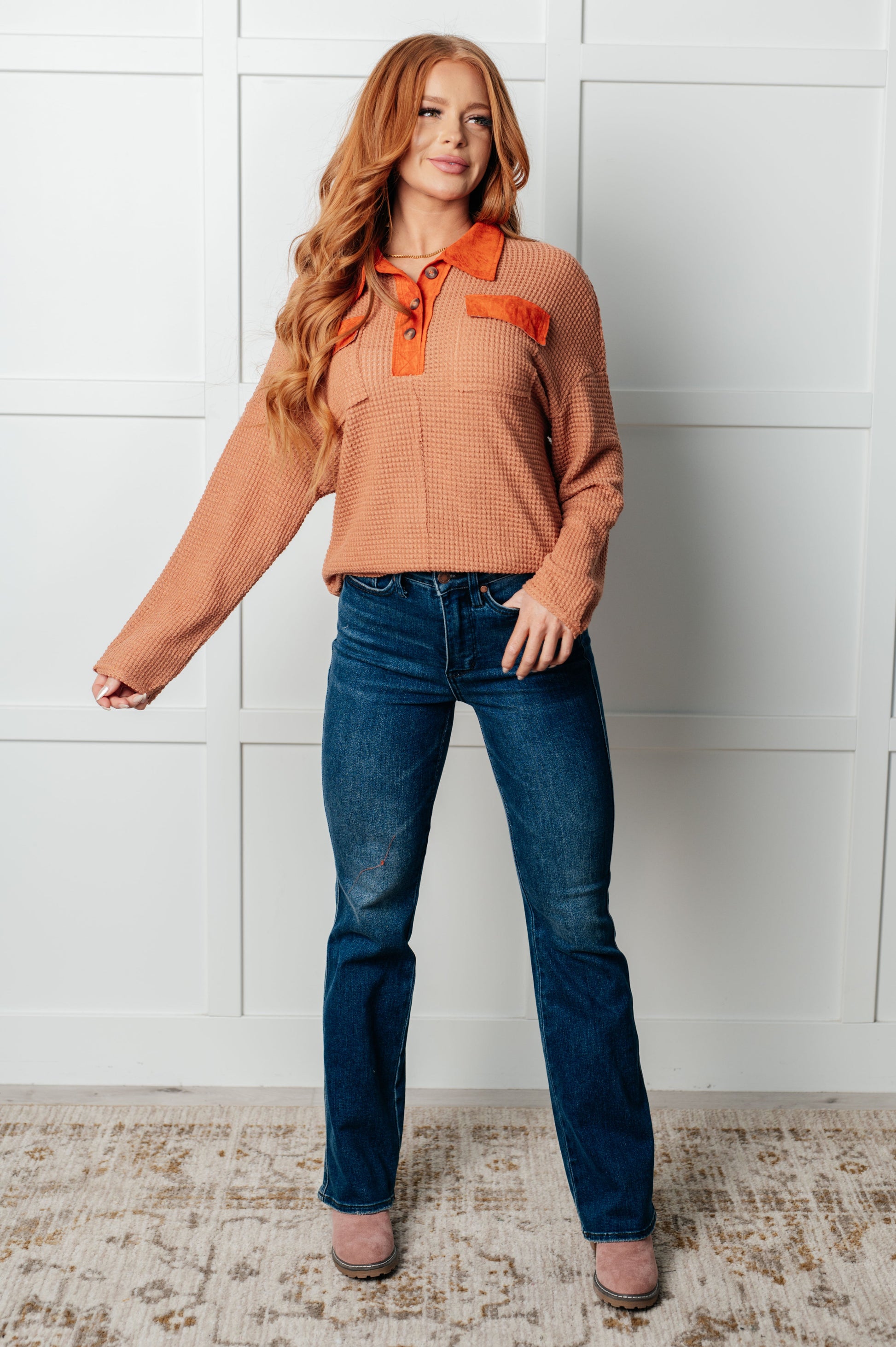 Woman wearing a brown sweater with orange accents and blue jeans against a white wall.