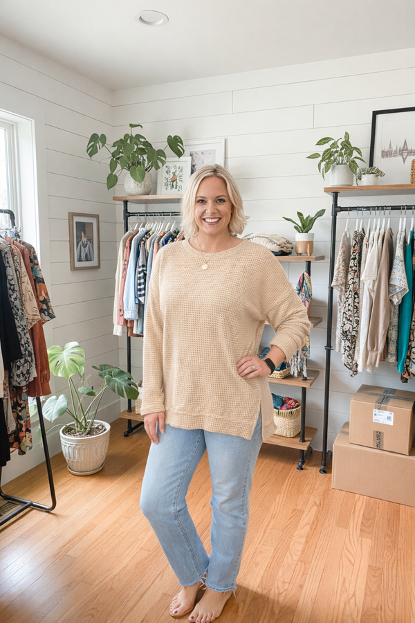 Woman standing in a clothing store wearing a beige sweater and light blue jeans.