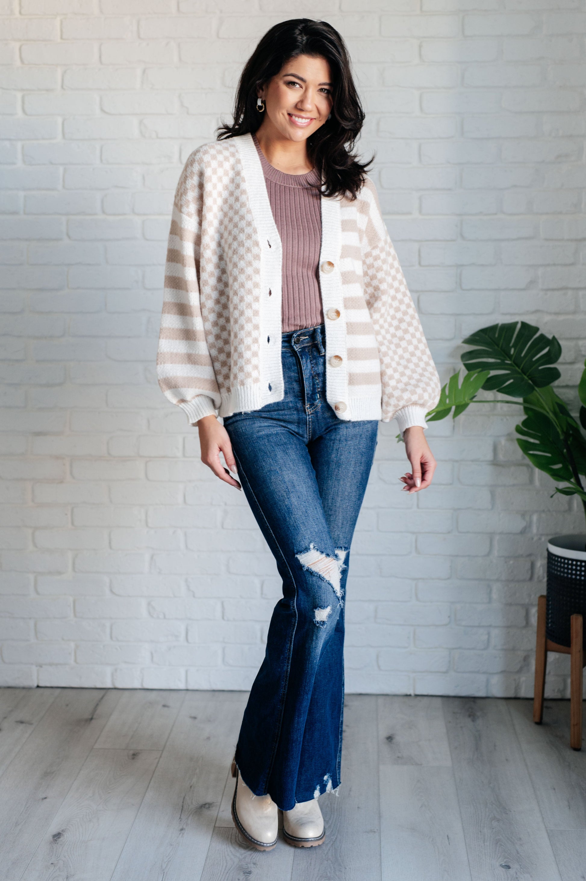 Woman wearing a white cardigan, pink top, and blue jeans standing against a white brick wall.