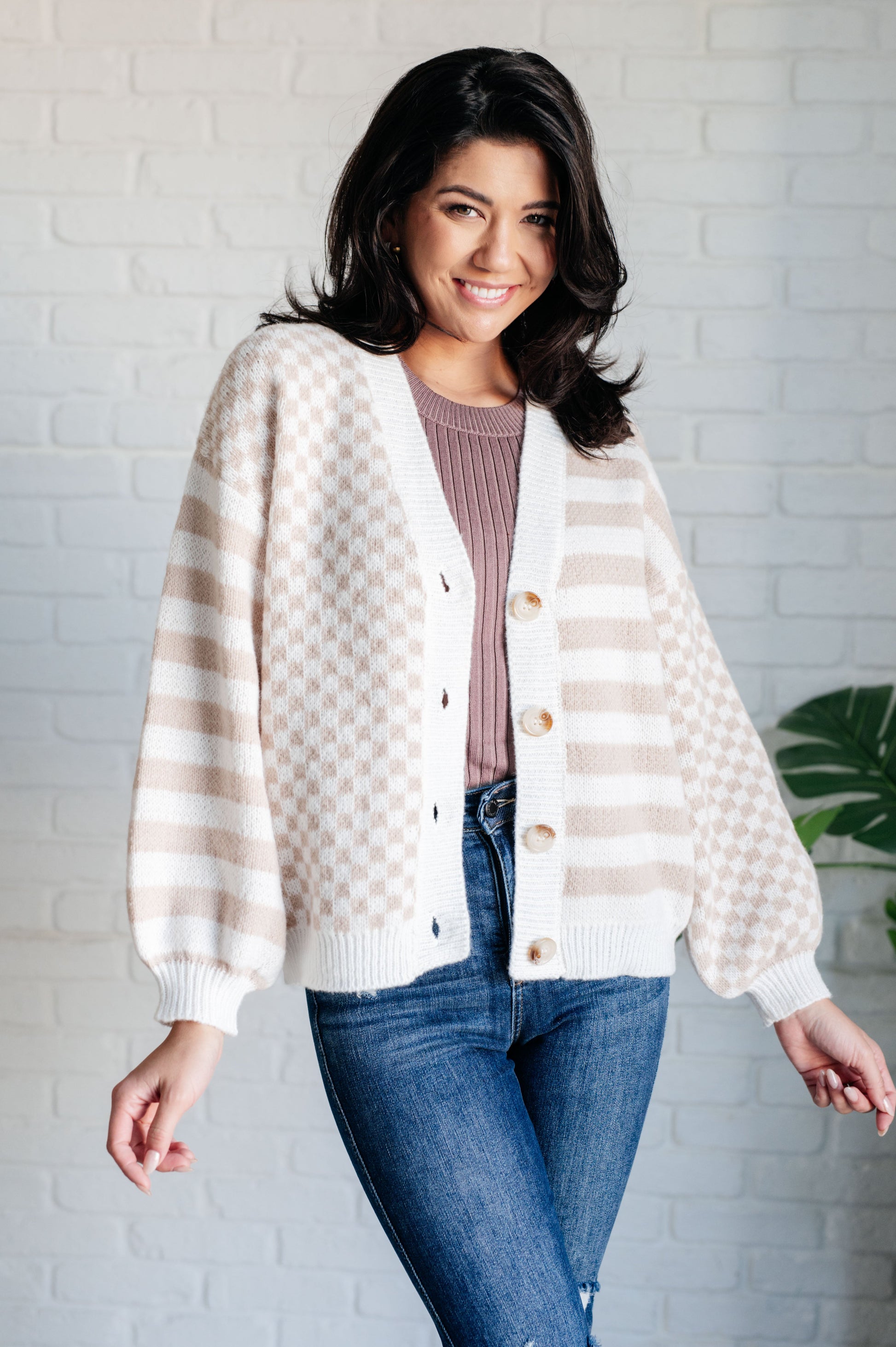 Woman wearing a striped cardigan over a sweater and jeans against a white brick wall.