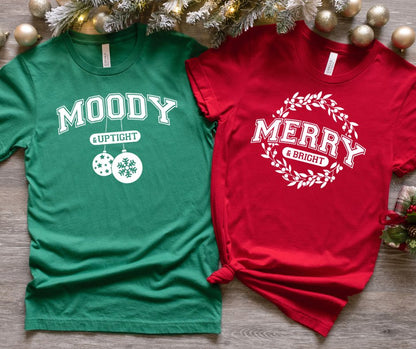 Two Christmas-themed t-shirts, one green and one red, on a wooden surface with decorative items.