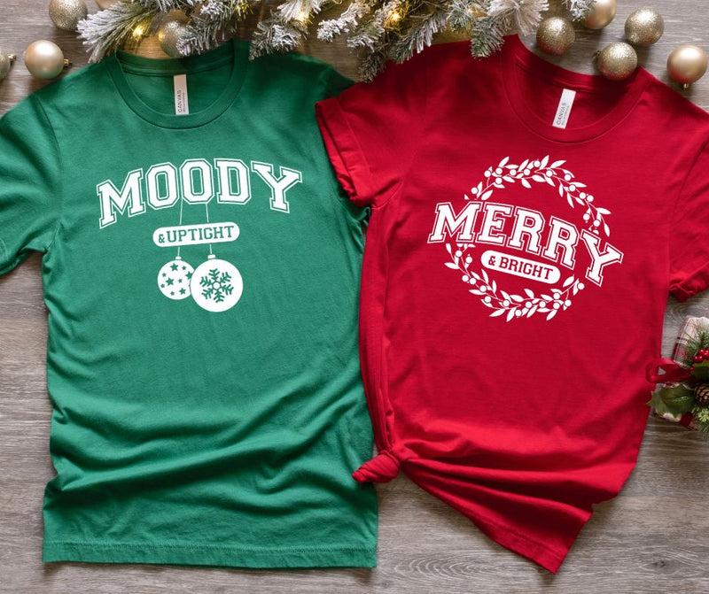 Two Christmas-themed t-shirts, one green and one red, on a wooden surface with decorative items.