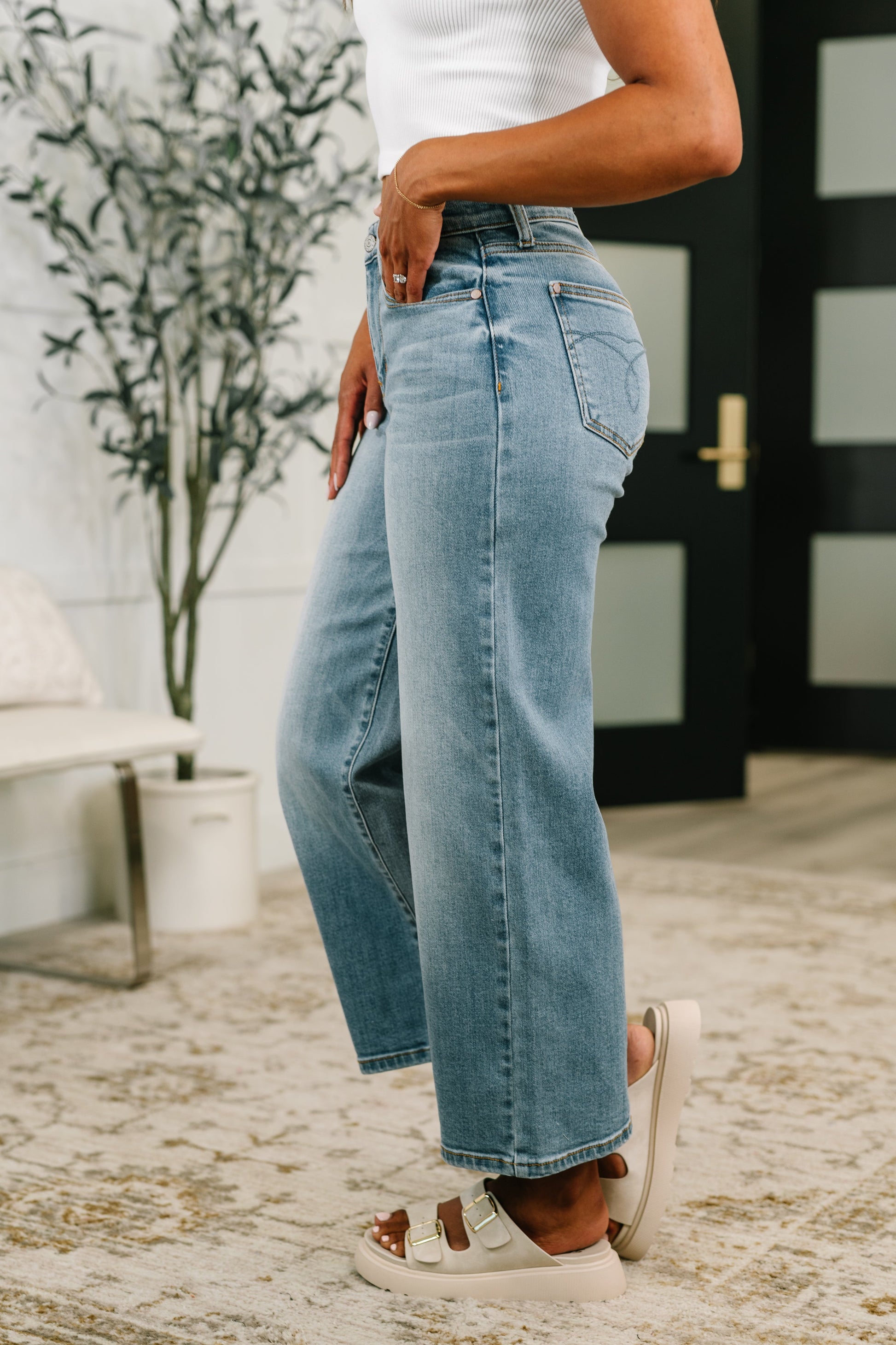 Person wearing light blue jeans standing indoors with a plant and door in the background