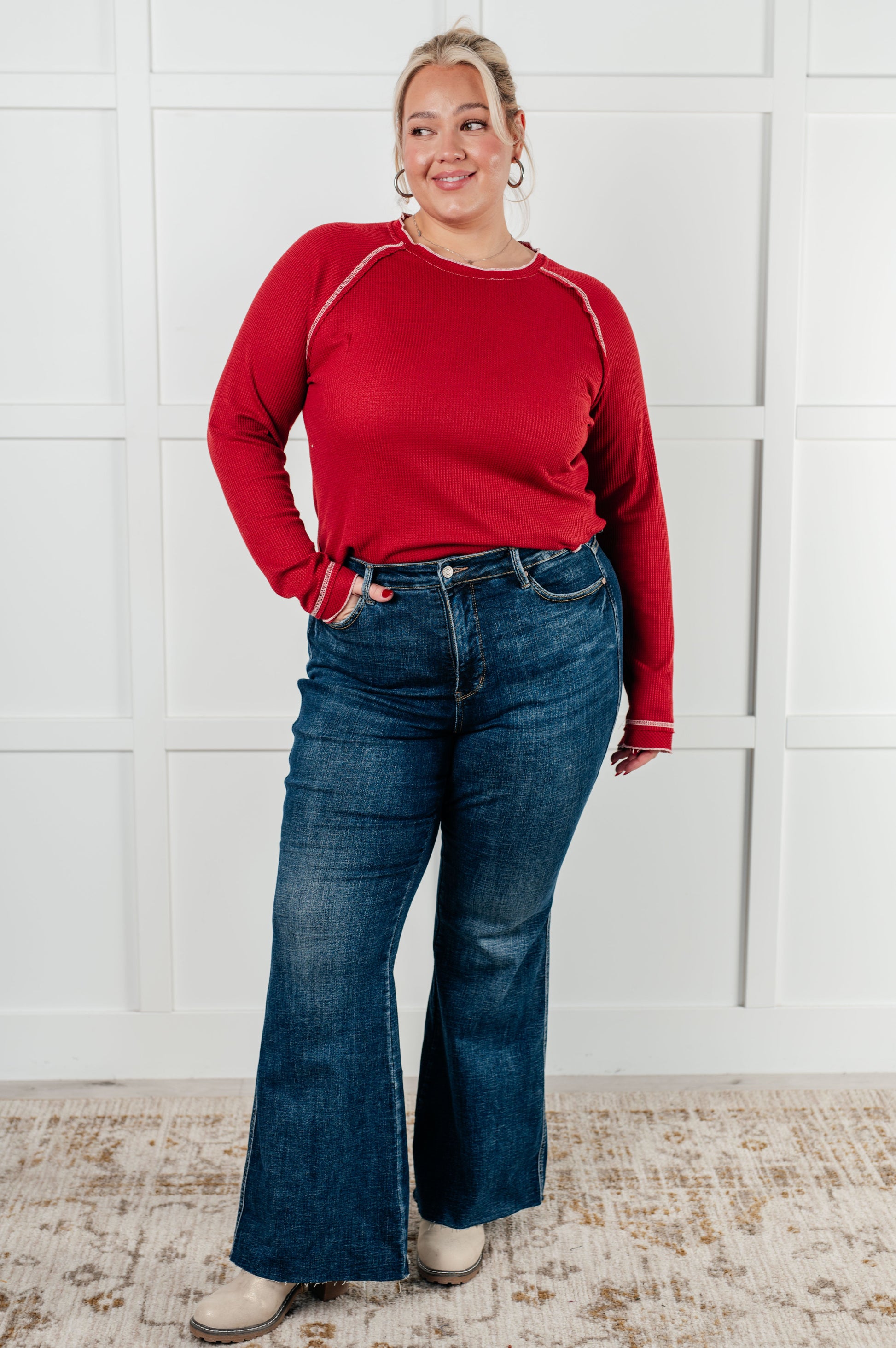 Person wearing a red long-sleeve shirt and blue jeans standing against a white brick wall.