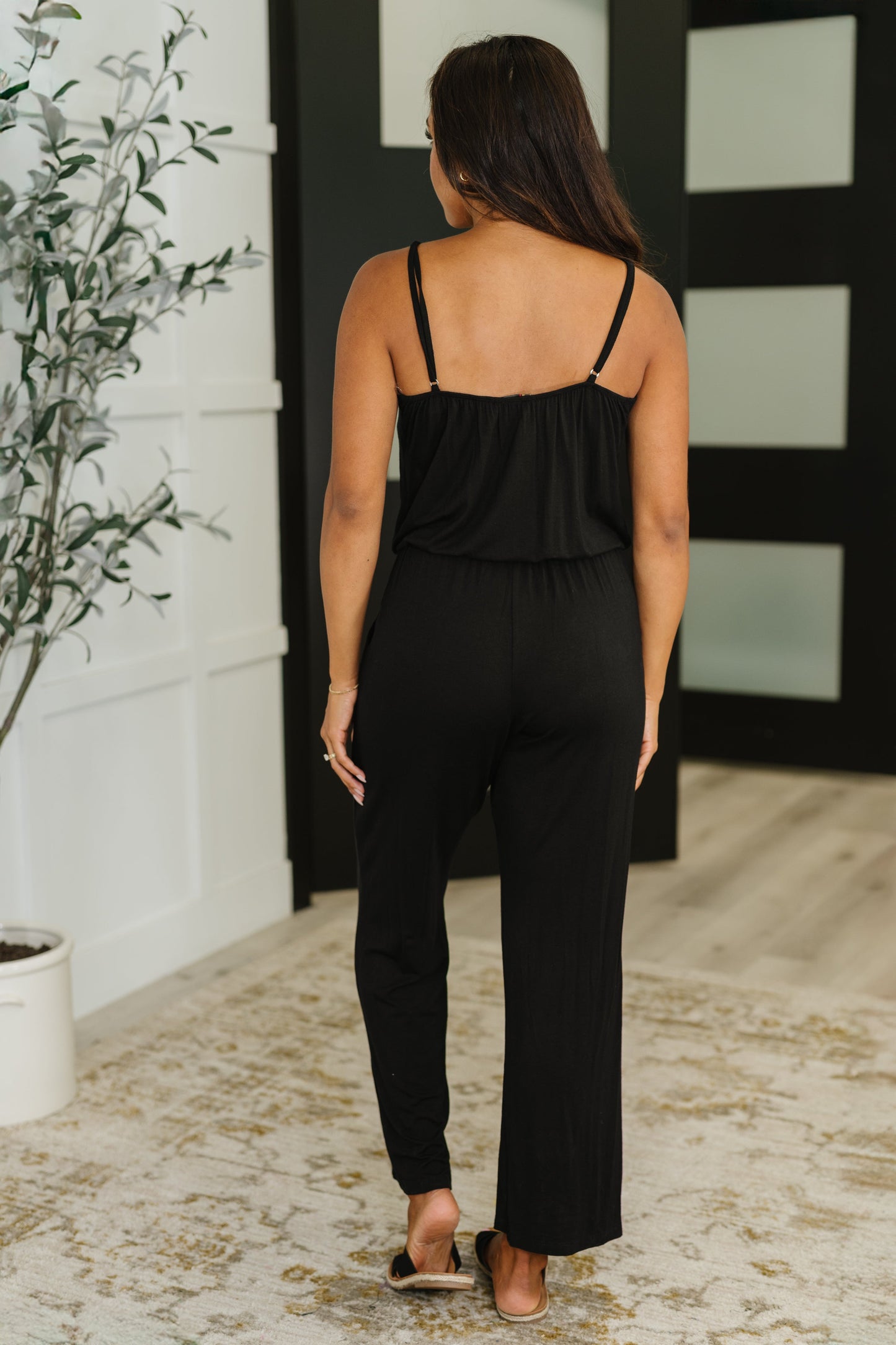 Woman wearing a black jumpsuit standing in a room with a plant and mirror.