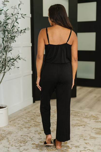Woman wearing a black jumpsuit standing in a room with a plant and mirror.
