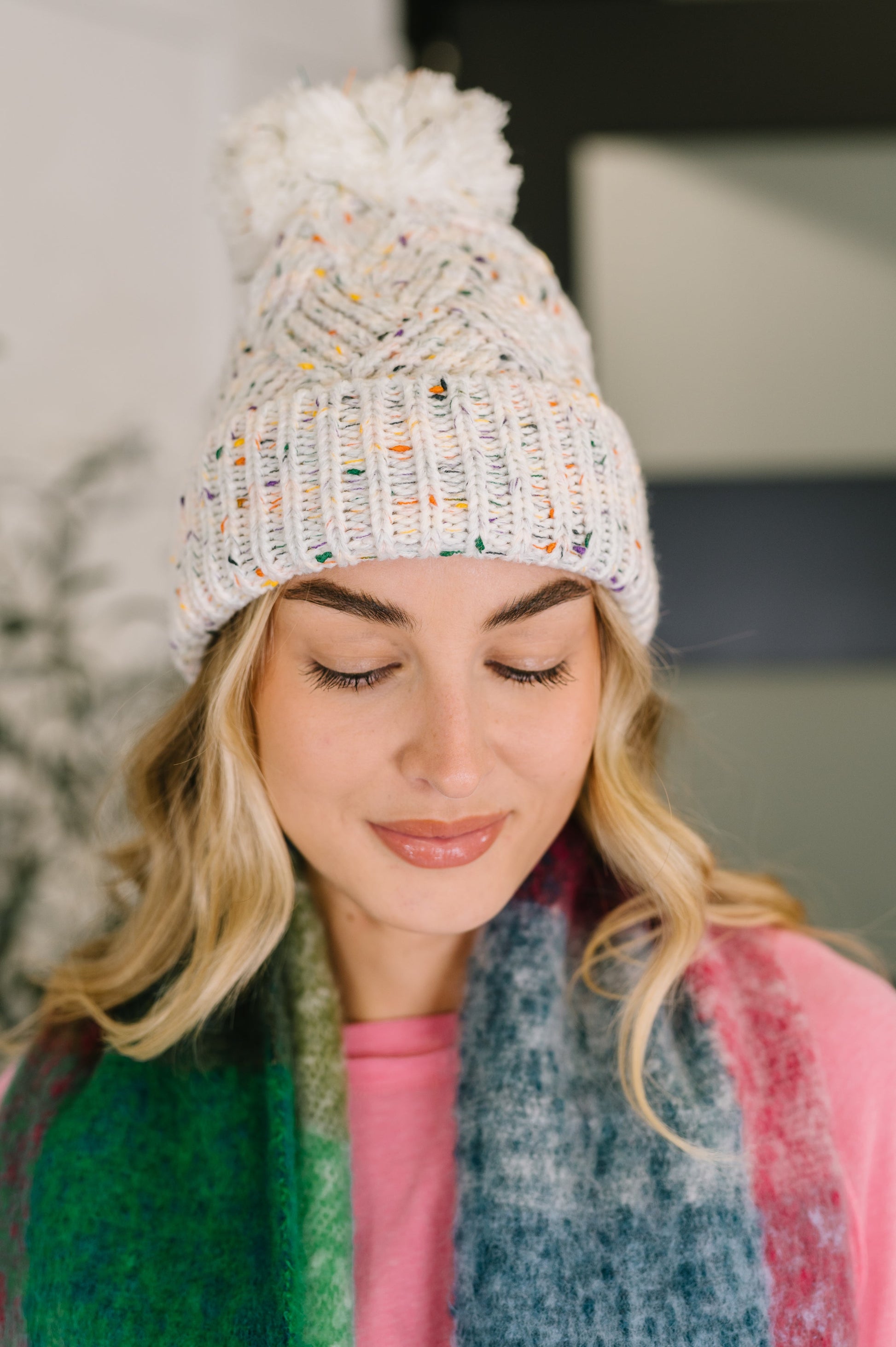 Woman wearing a white knit beanie with colorful accents and a fluffy pom-pom, looking down.