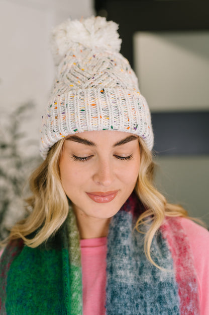 Woman wearing a white knit beanie with colorful accents and a fluffy pom-pom, looking down.