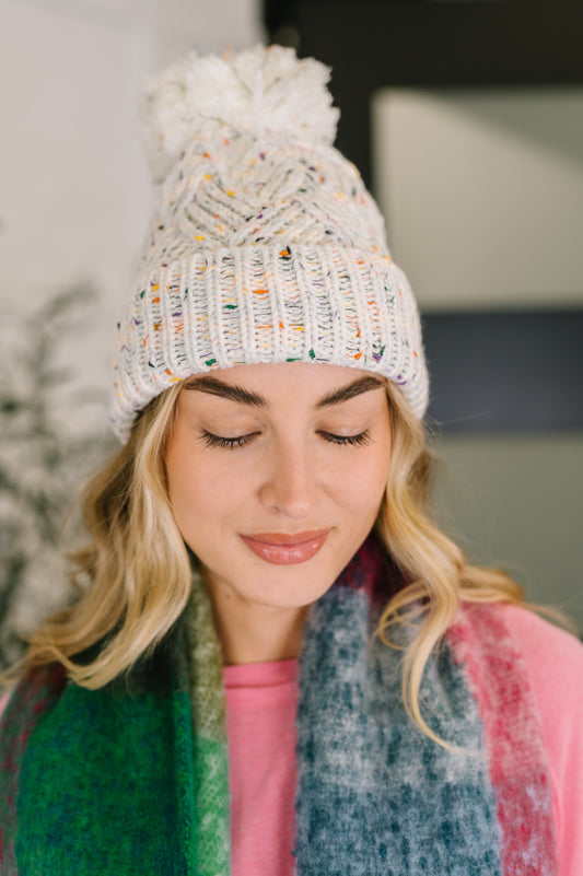 Woman wearing a white knit beanie with colorful accents and a fluffy pom-pom, looking down.