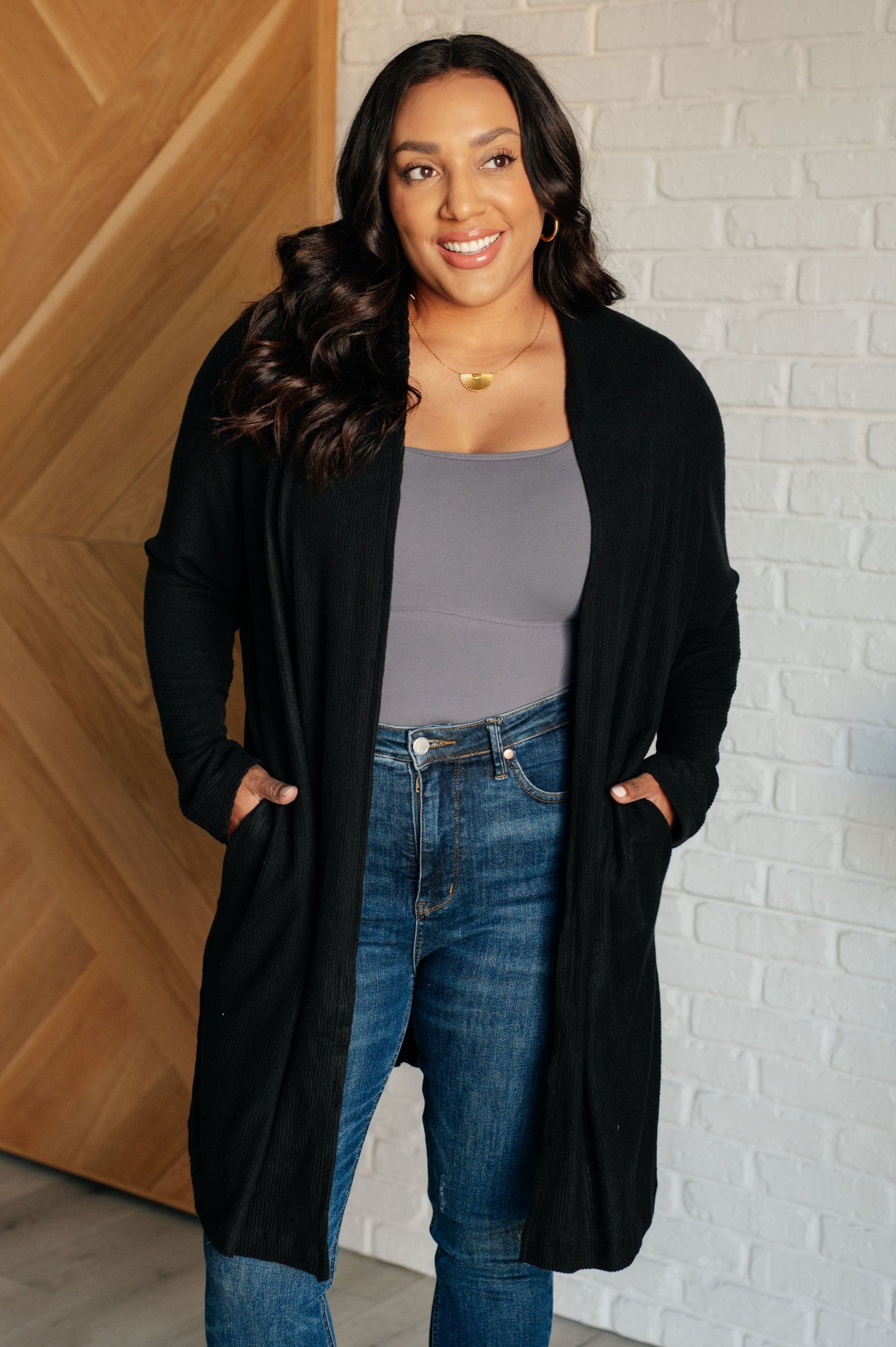 Woman wearing a black cardigan, gray top, and blue jeans against a brick wall.