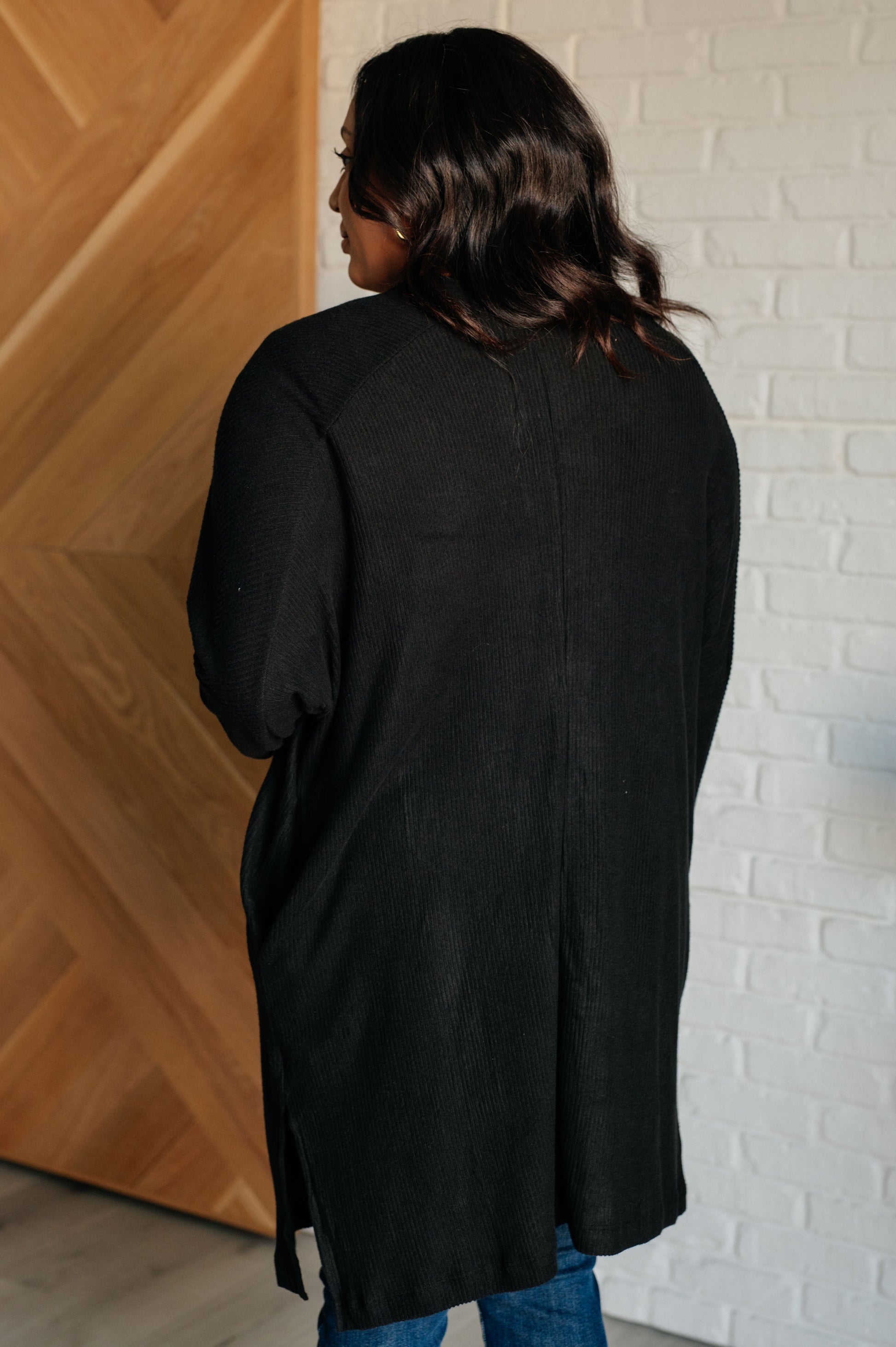 Person wearing a black cardigan standing against a white brick wall.