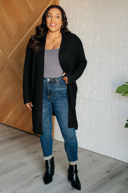 Woman wearing a black cardigan, gray top, blue jeans, and black boots standing against a white brick wall.
