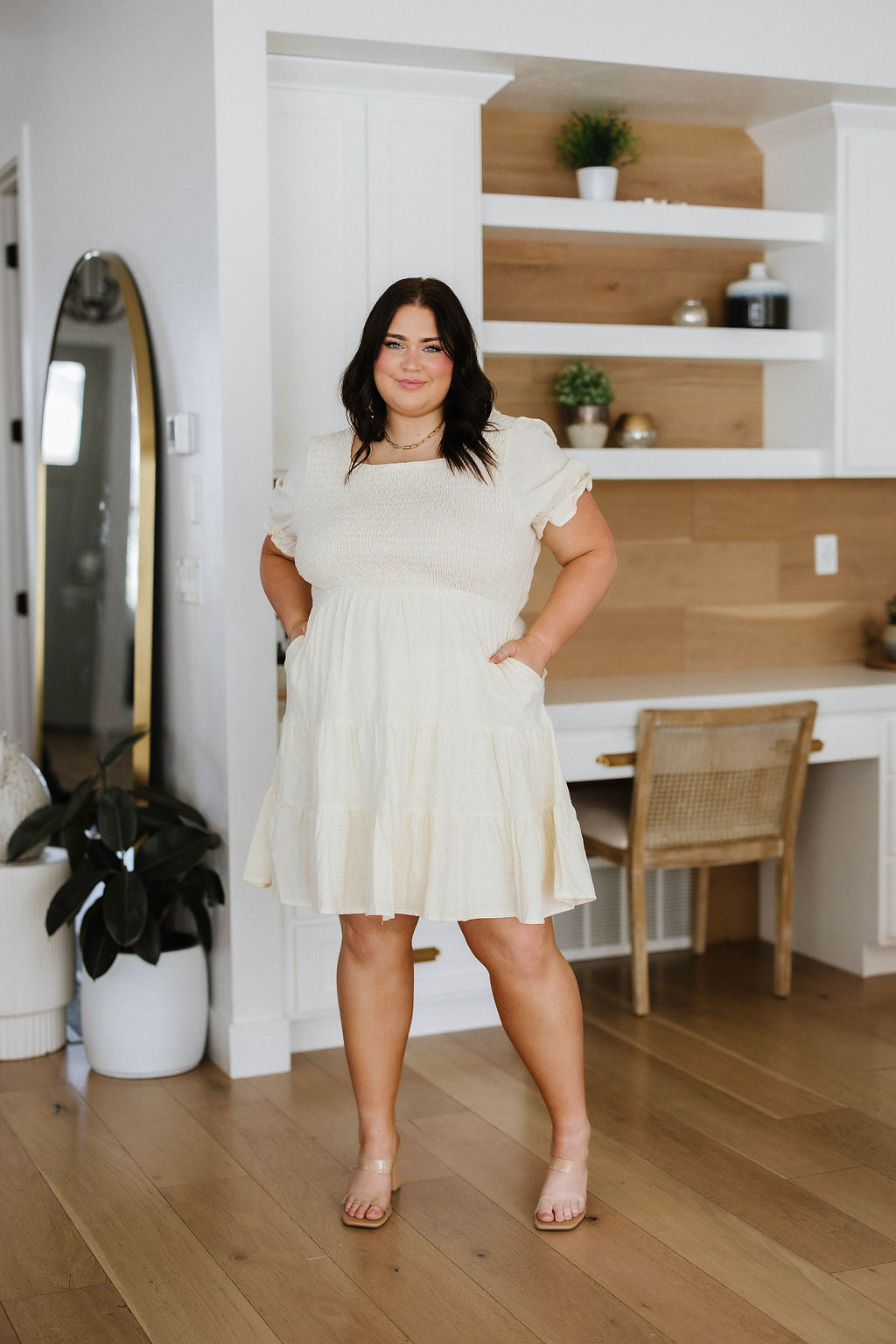 Woman in a white dress standing in a modern interior setting with wooden floors and shelves.