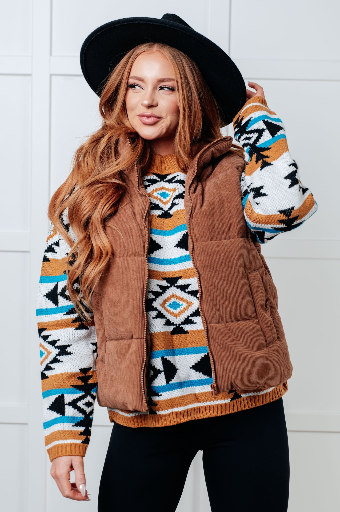 Woman wearing a patterned sweater with a brown vest against a white tiled wall.