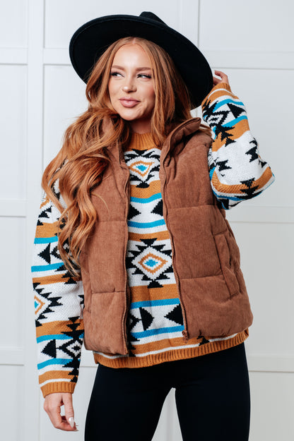 Woman wearing a patterned sweater with a brown vest against a white tiled wall.