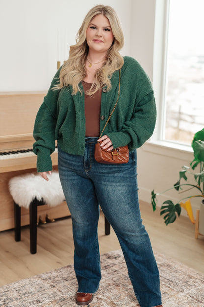 Woman wearing a green cardigan, brown top, and blue jeans in a room with a piano and window.