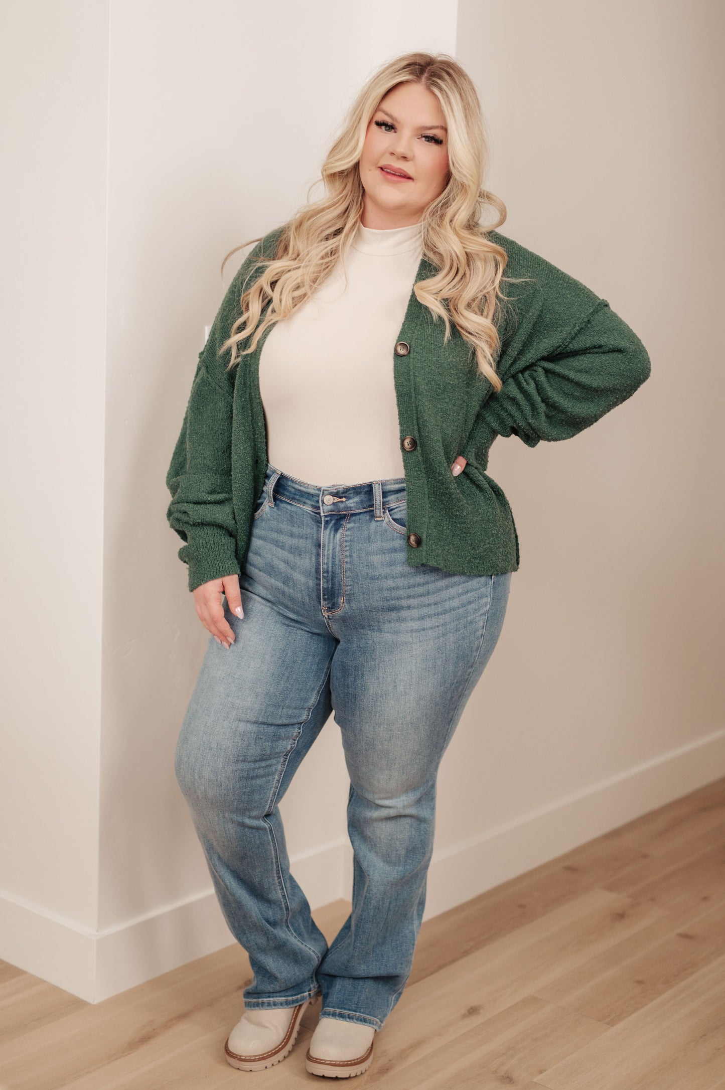 Woman wearing a green cardigan, white top, and blue jeans against a plain wall.