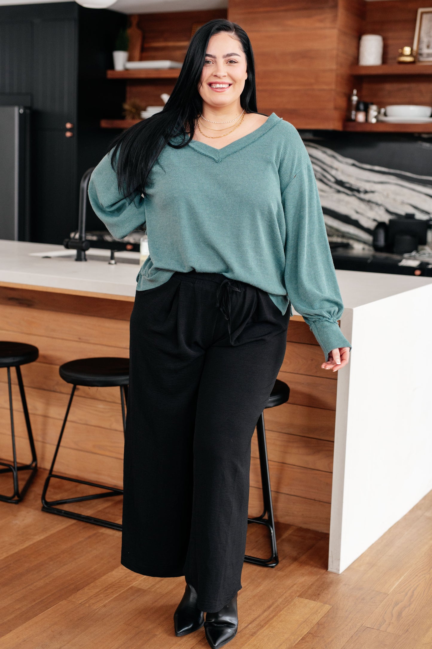 Woman standing in a kitchen wearing a teal sweater and black pants.