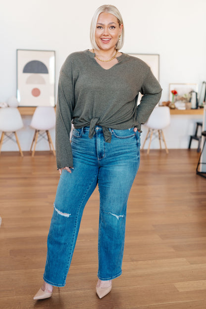 Woman wearing a gray long-sleeve top and blue jeans standing in a room with wooden flooring and white chairs.