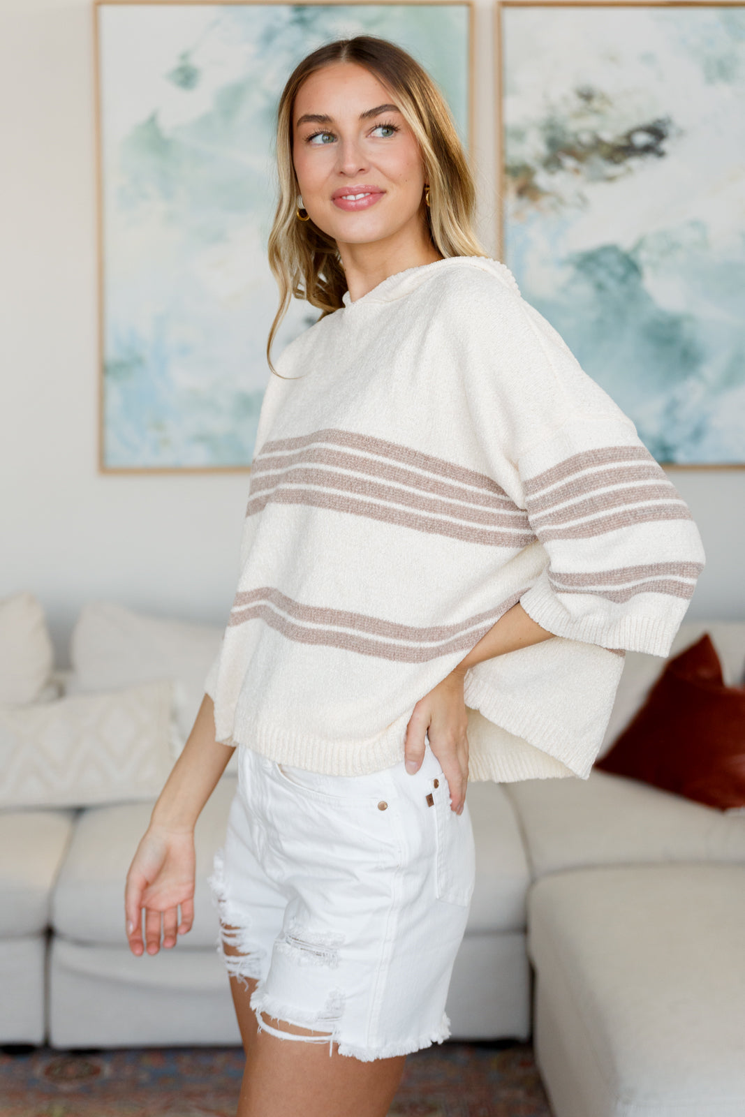 Woman wearing a white sweater with brown stripes and white shorts in a living room.