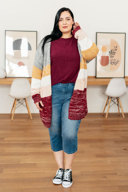 Woman wearing a colorful cardigan, maroon top, and blue jeans in a room with wooden flooring and framed pictures on the wall.