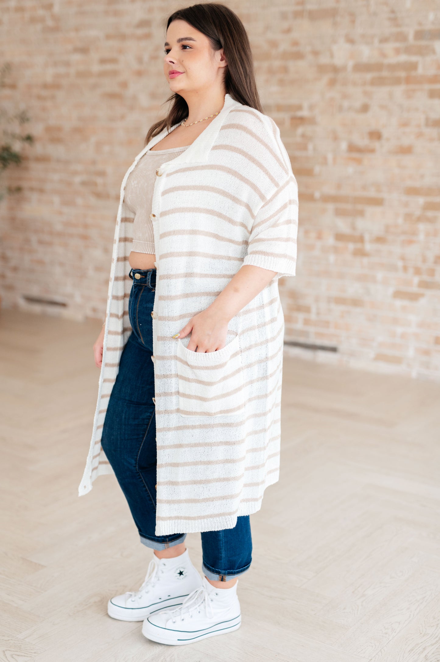 Woman wearing a long, light-colored striped cardigan over a beige top and blue jeans, standing against a brick wall.