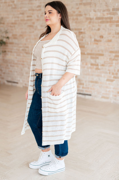 Woman wearing a long, light-colored striped cardigan over a beige top and blue jeans, standing against a brick wall.
