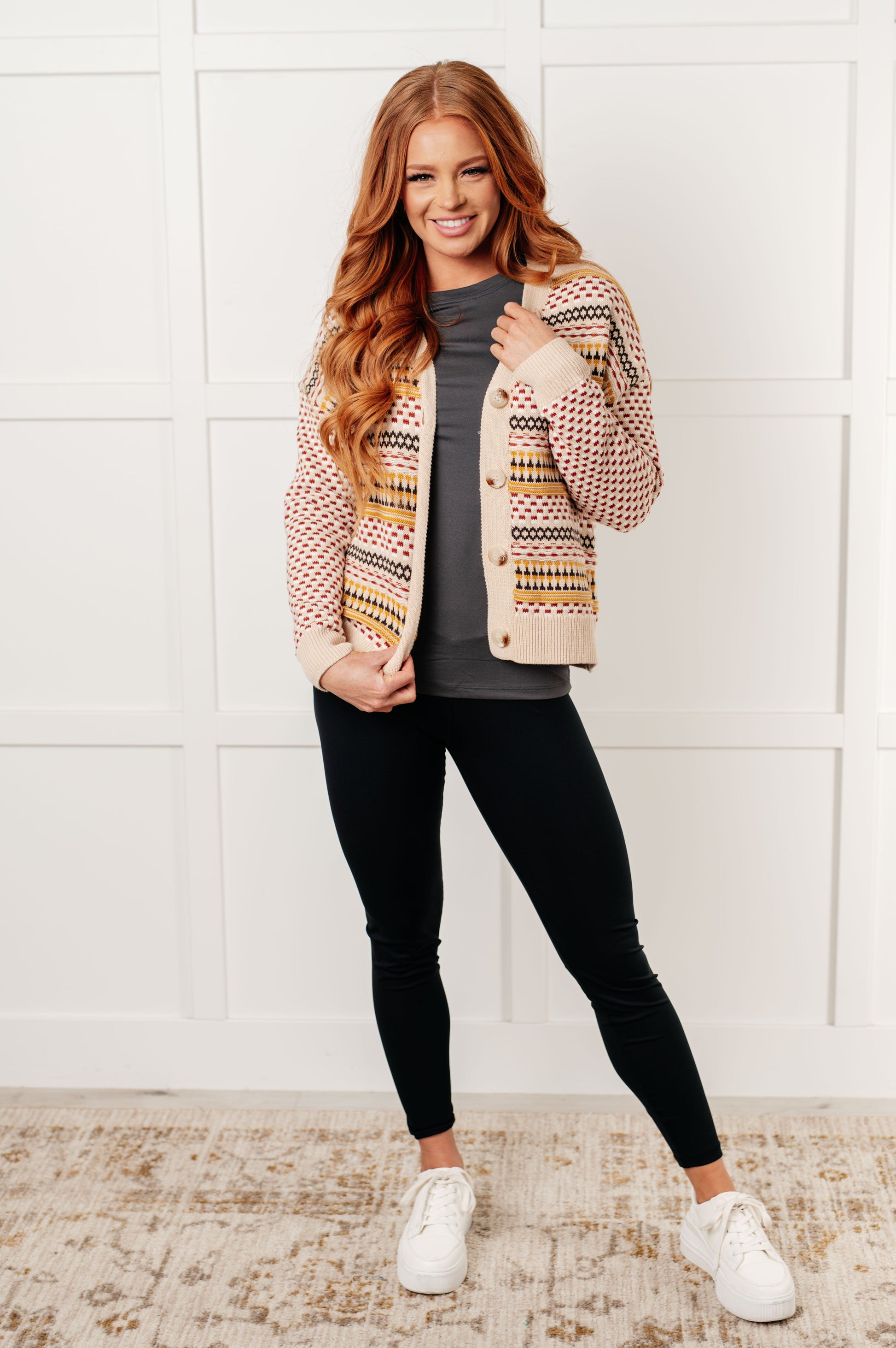 Woman wearing a patterned cardigan, gray shirt, black leggings, and white sneakers against a white wall.