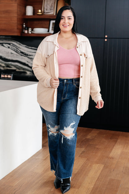 Woman standing in a kitchen wearing a beige jacket, pink top, and blue jeans.
