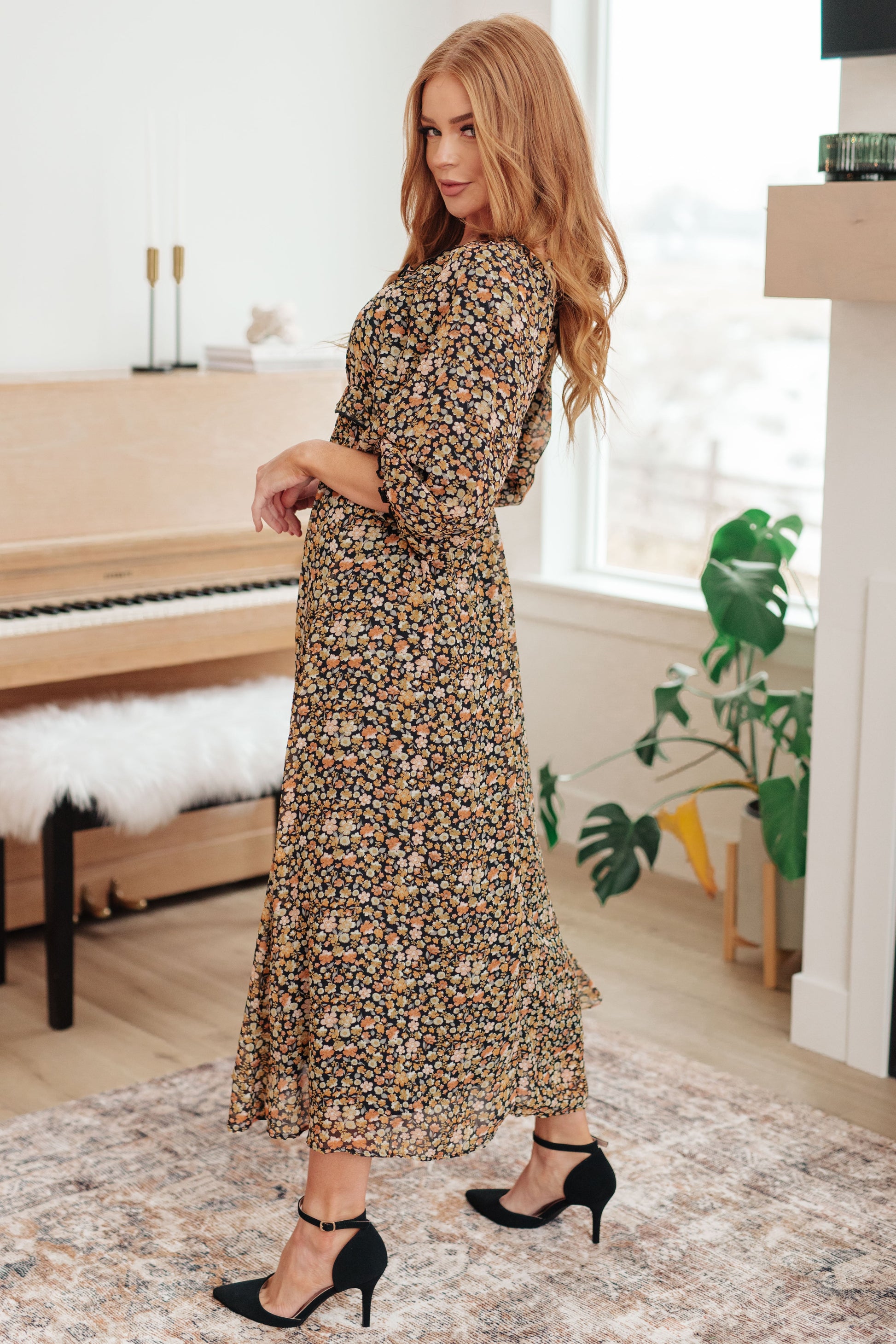 Woman in a floral dress standing in a room with a piano and plants.