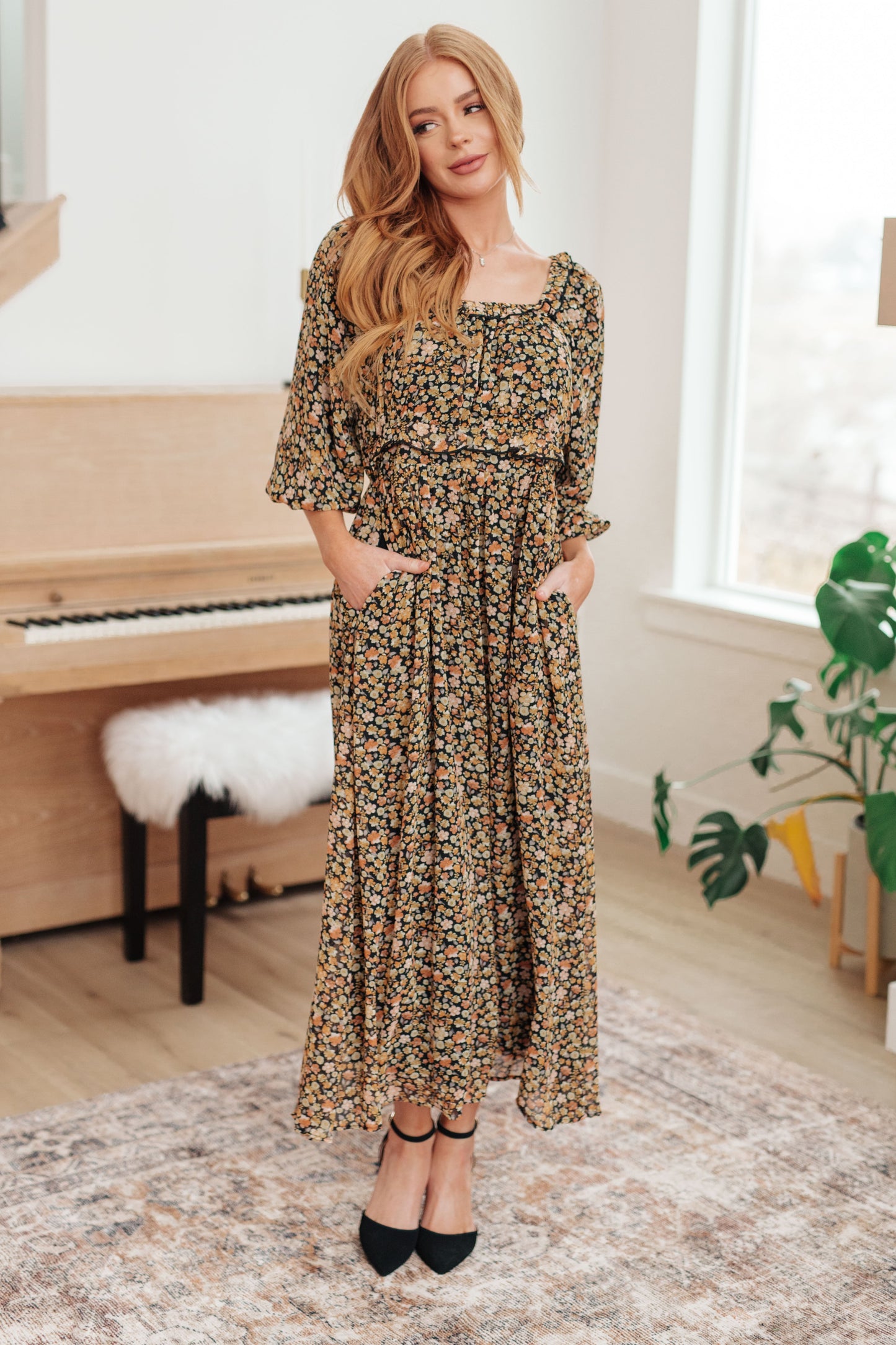 Woman in a floral dress standing in a room with a piano and plant.