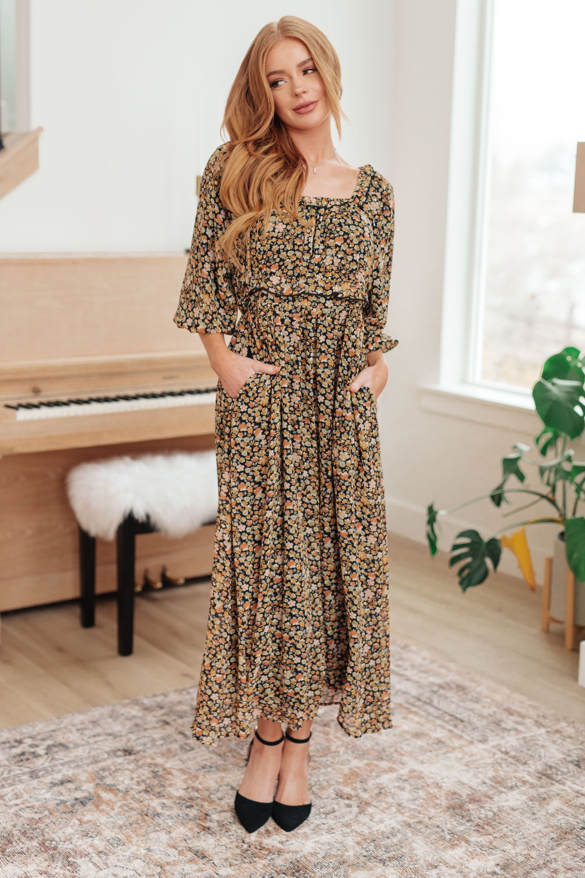 Woman in a floral dress standing in a room with a piano and plant.