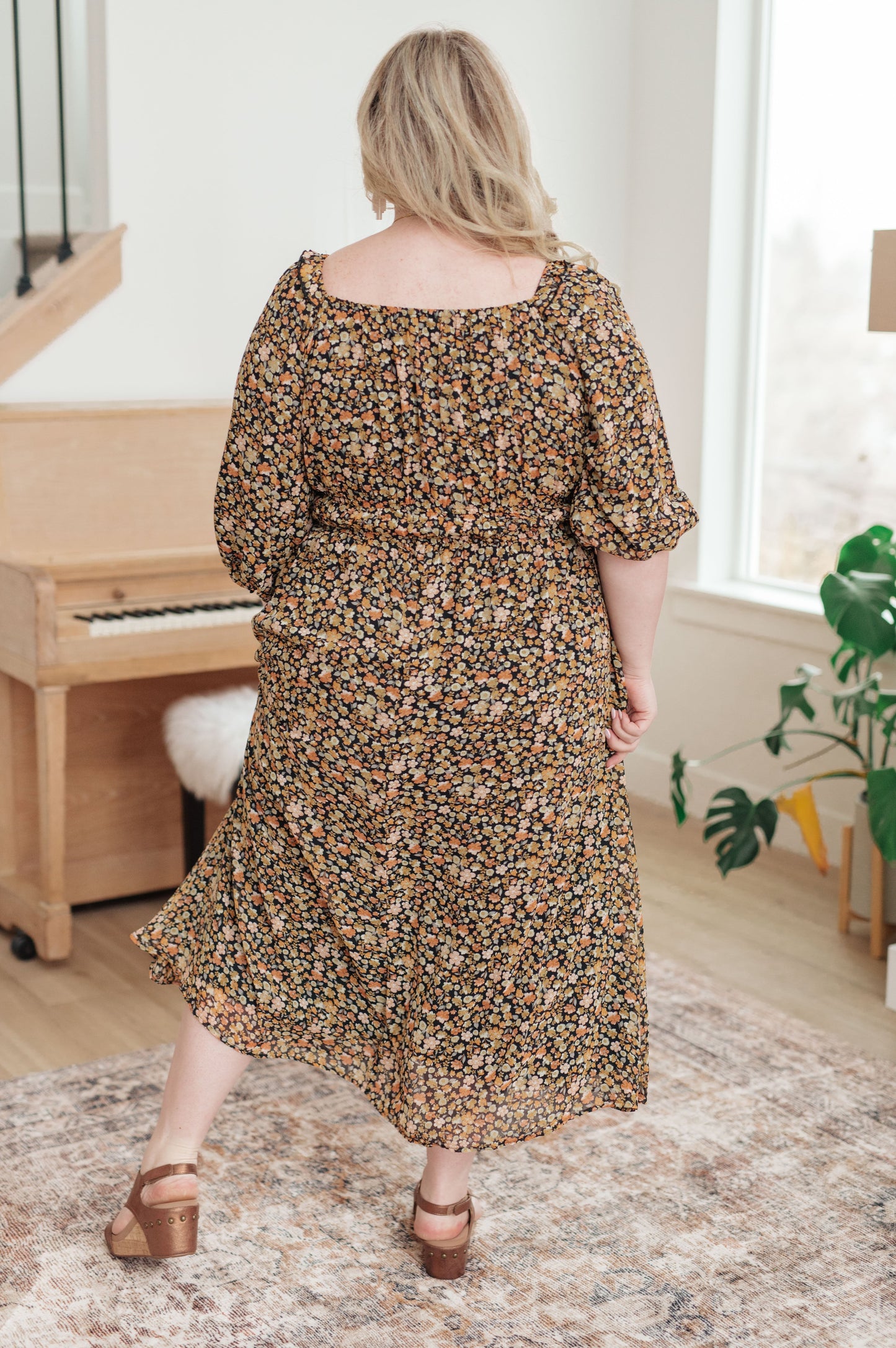 Woman in a floral dress standing in a room with a piano and window.