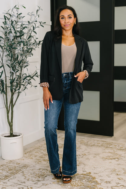 Woman in black blazer and blue jeans standing indoors with a plant and decorative wall in the background