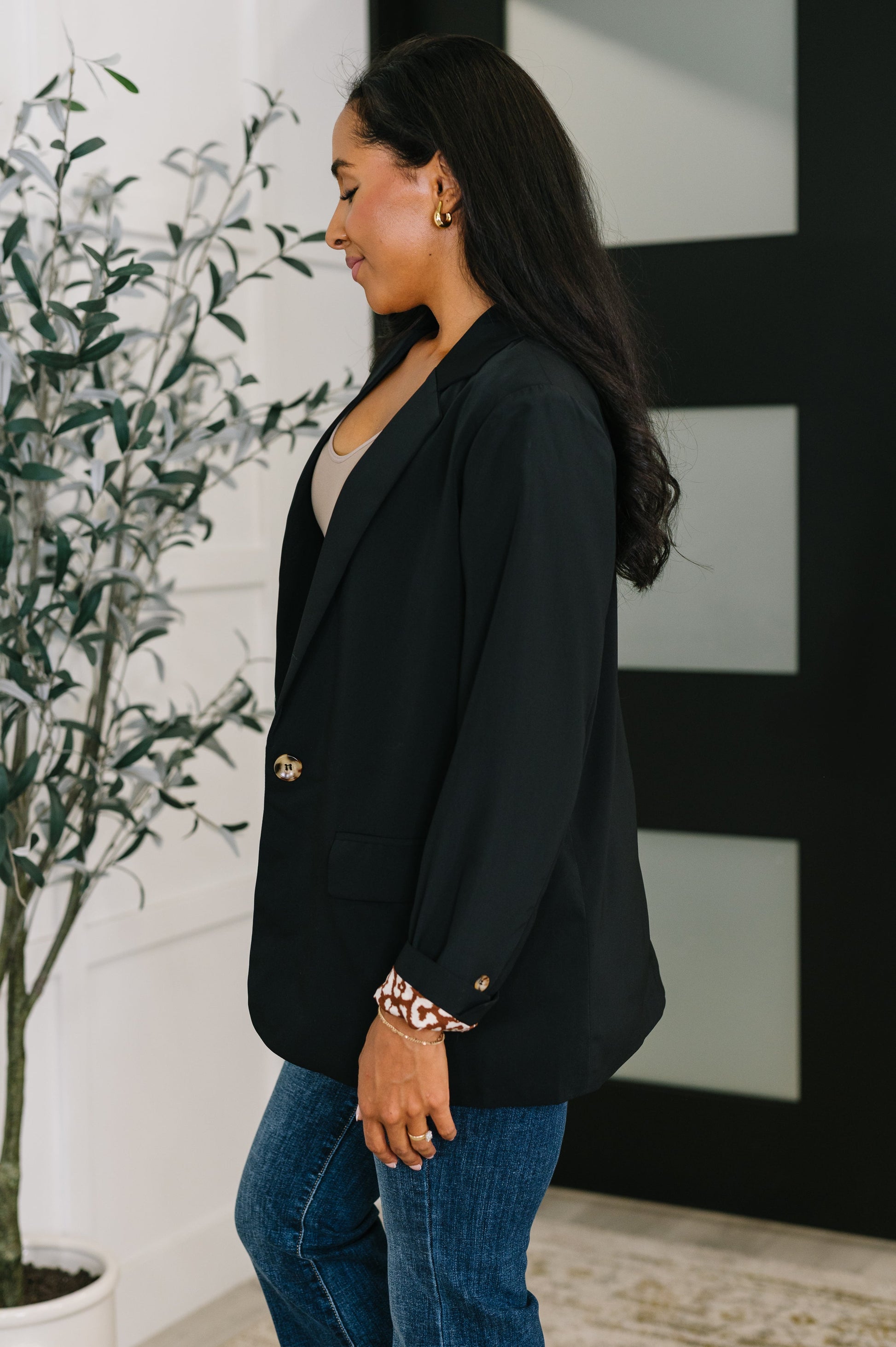 Woman wearing a black blazer and blue jeans standing indoors with a plant and abstract art in the background.