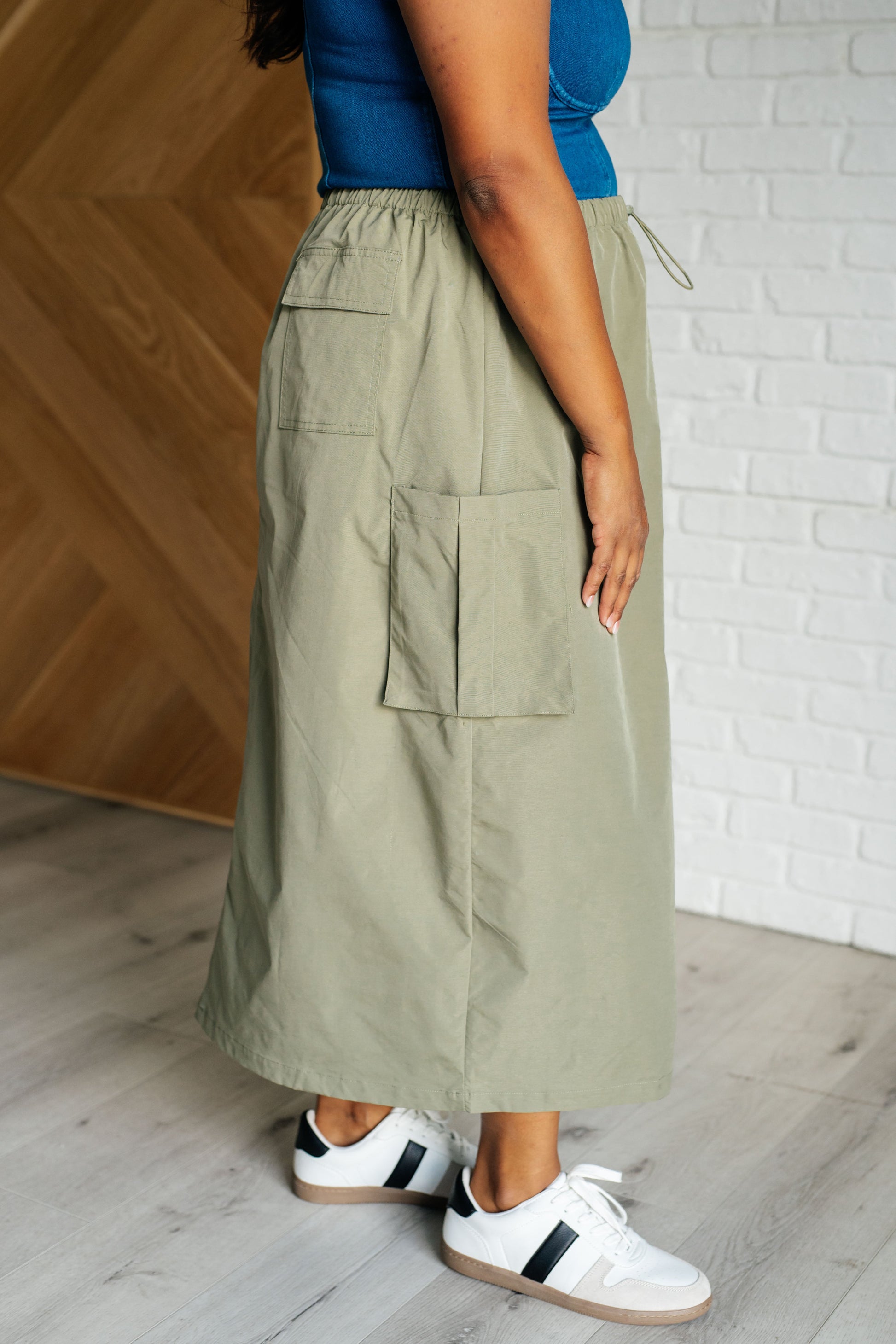 Person wearing a long olive green skirt with pockets, standing against a wooden and white brick wall.