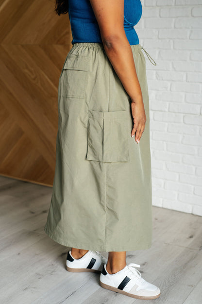 Person wearing a long olive green skirt with pockets, standing against a wooden and white brick wall.