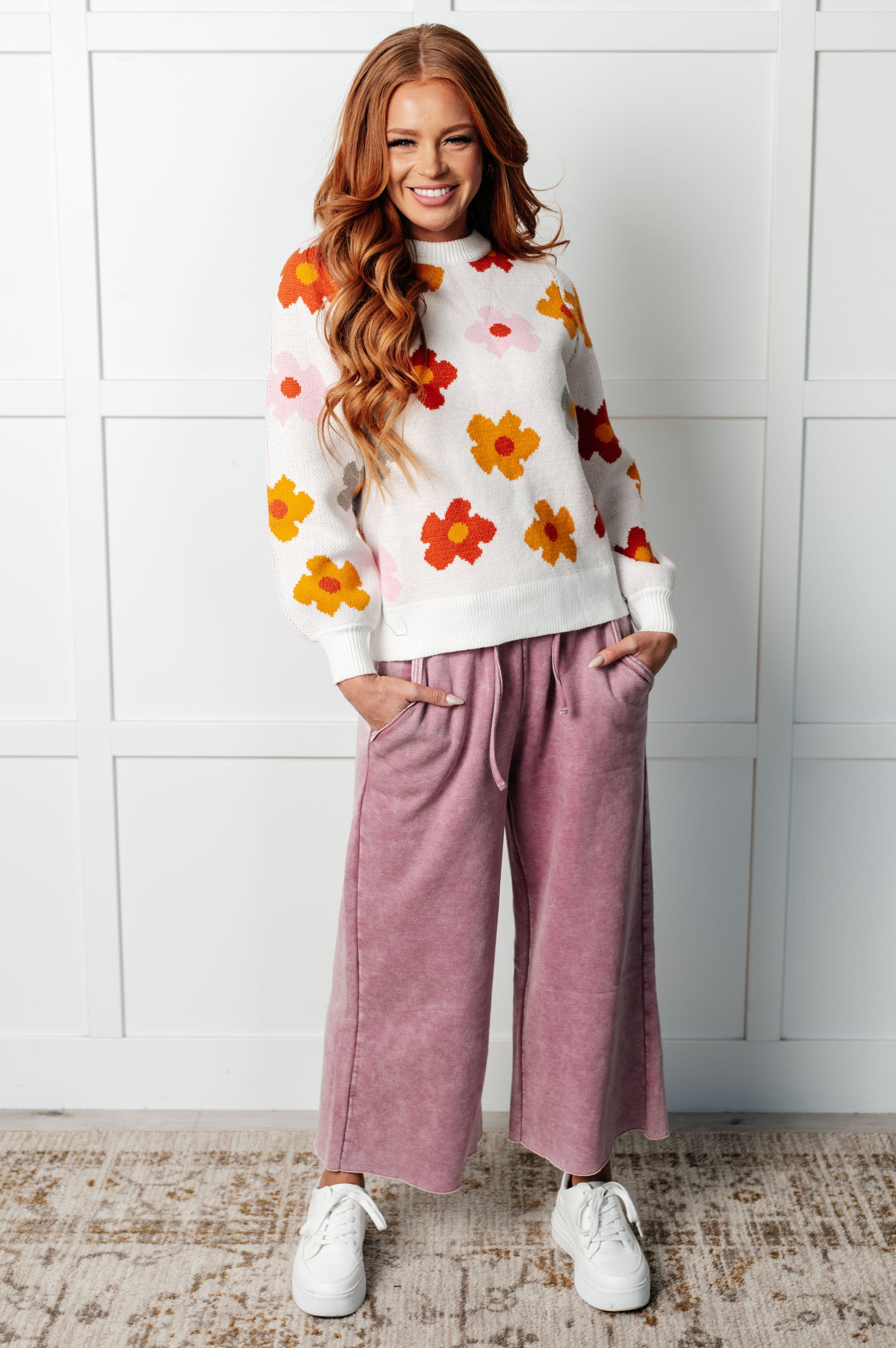 Woman wearing a floral sweater and pink pants against a white wall.