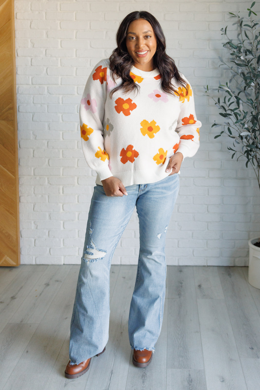 Woman wearing a floral sweater and jeans standing in a room with a white brick wall and plant.